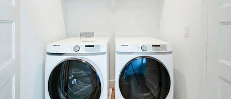 Two white Samsung front-loading washing machines in a laundry room with a wooden floor and white walls.