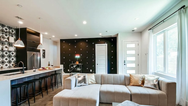A modern living room with a sofa, ottoman, and a coffee table. There is a kitchen area with a refrigerator, sink, and bar stools. The room has a black wall with musical notes and a white door.