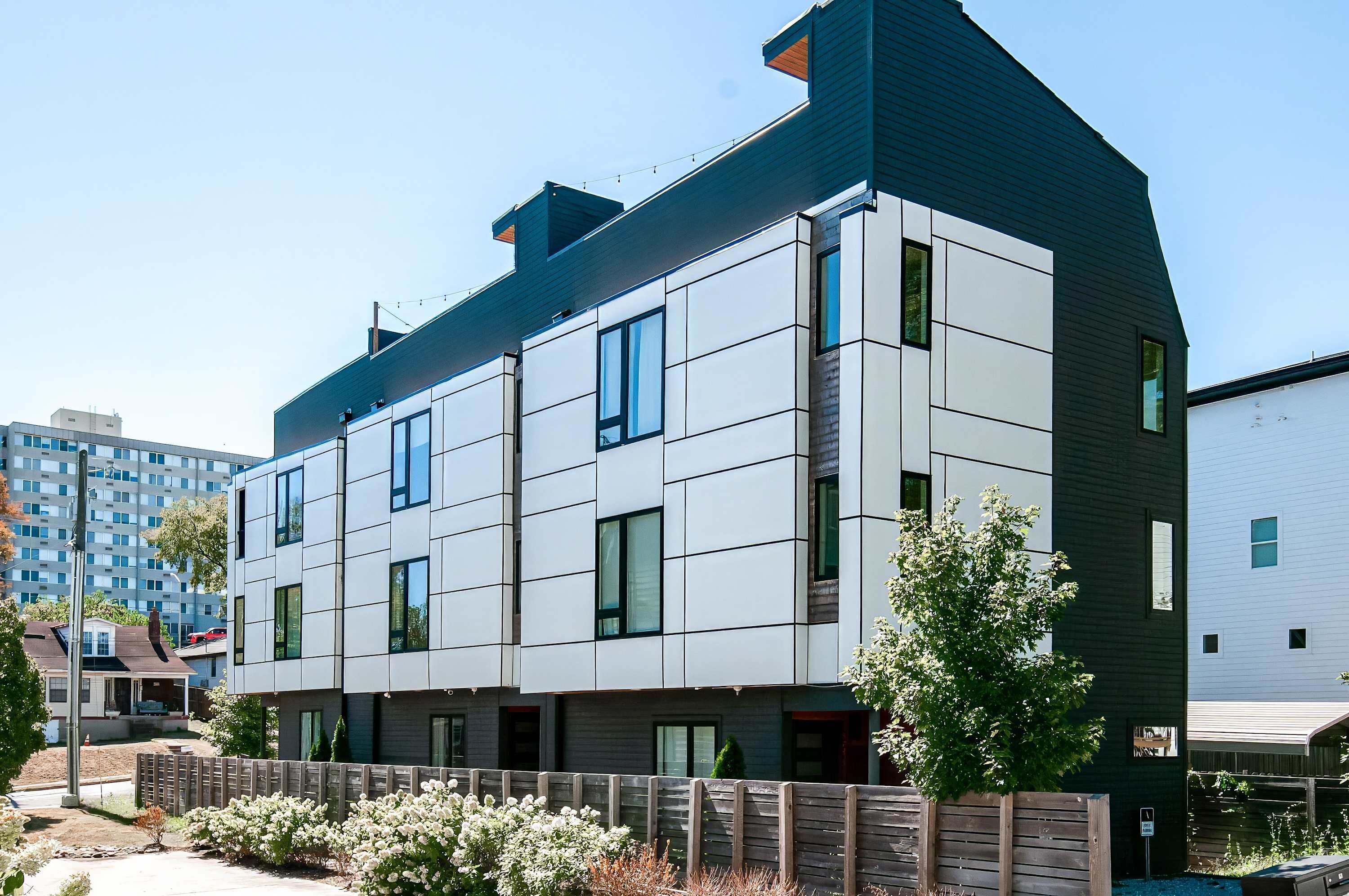 A modern multi-story residential building with a flat roof, large windows, and a wooden fence in the foreground.
