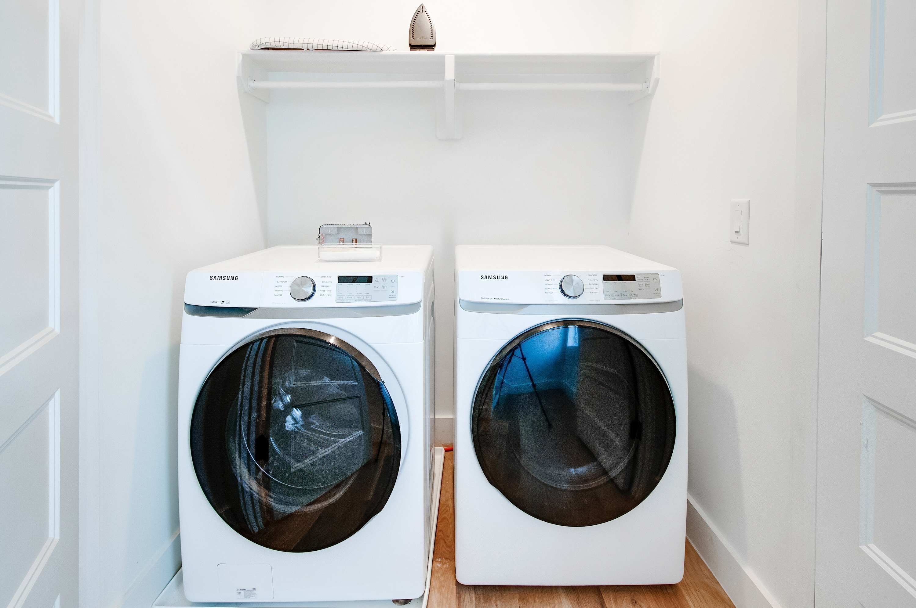 Dos lavadoras Samsung blancas de carga frontal en un cuarto de lavado con paredes blancas y piso de madera.