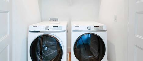 Dos lavadoras Samsung blancas de carga frontal en un cuarto de lavado con paredes blancas y piso de madera.