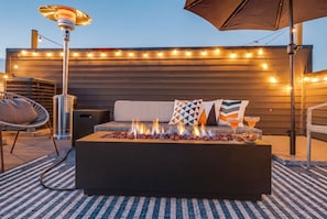 An outdoor seating area with a fire feature, string lights, and a table with drinks.