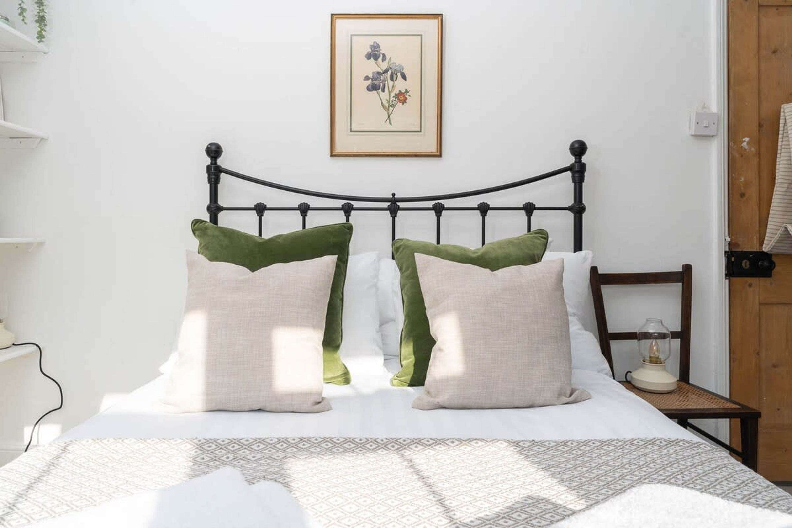 Een net opgemaakt bed met een bedsprei met een patroon, twee groene kussens, en een bloemig kunstwerk in een lijst aan de muur.