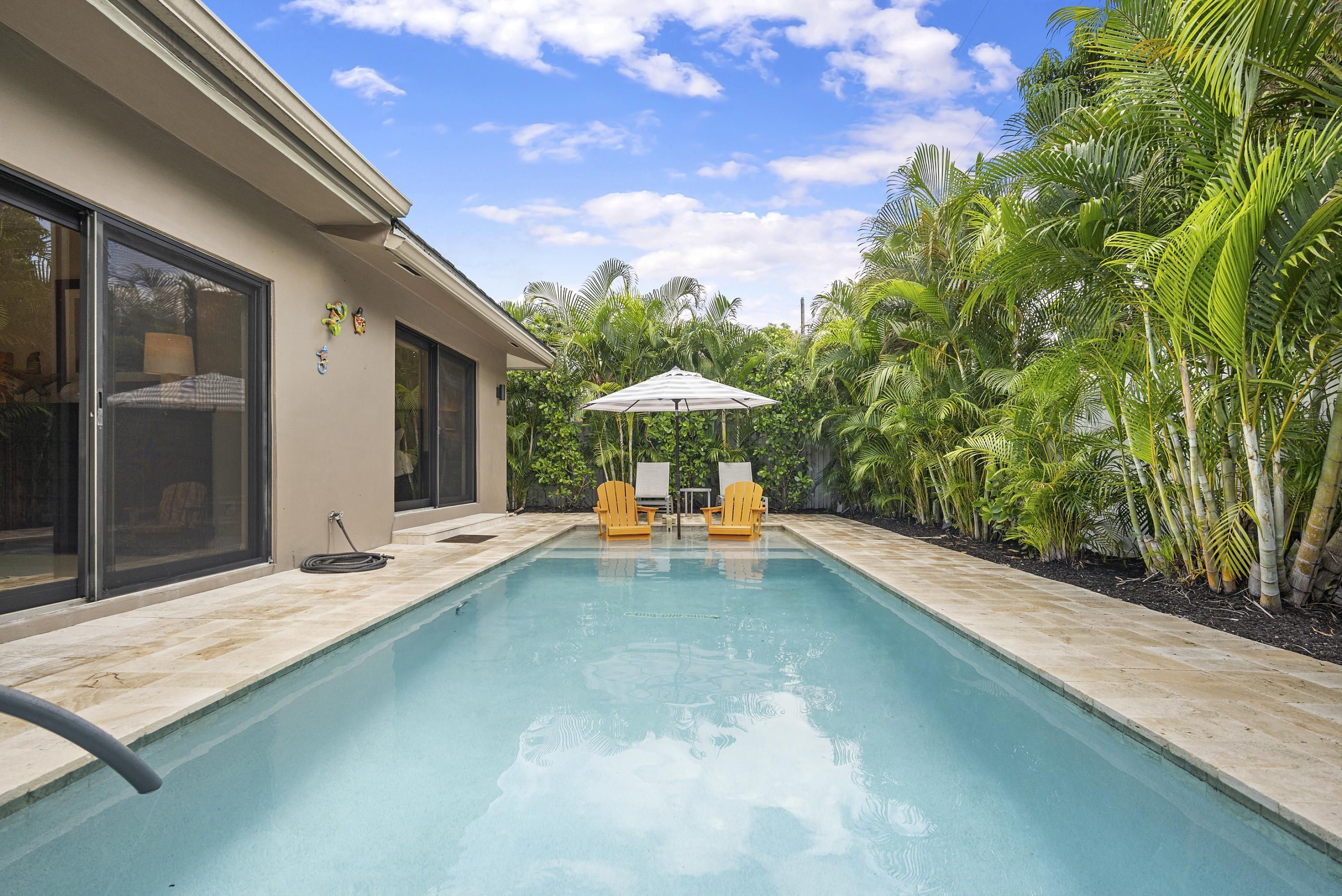 A swimming pool with two yellow chairs and a white umbrella, surrounded by lush greenery and a house with large windows.