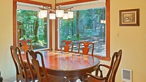 A dining area with a round table and six chairs, large windows with a forest view, and a framed picture on the wall. - Lindgren Hause in Enumclaw (Enumclaw)