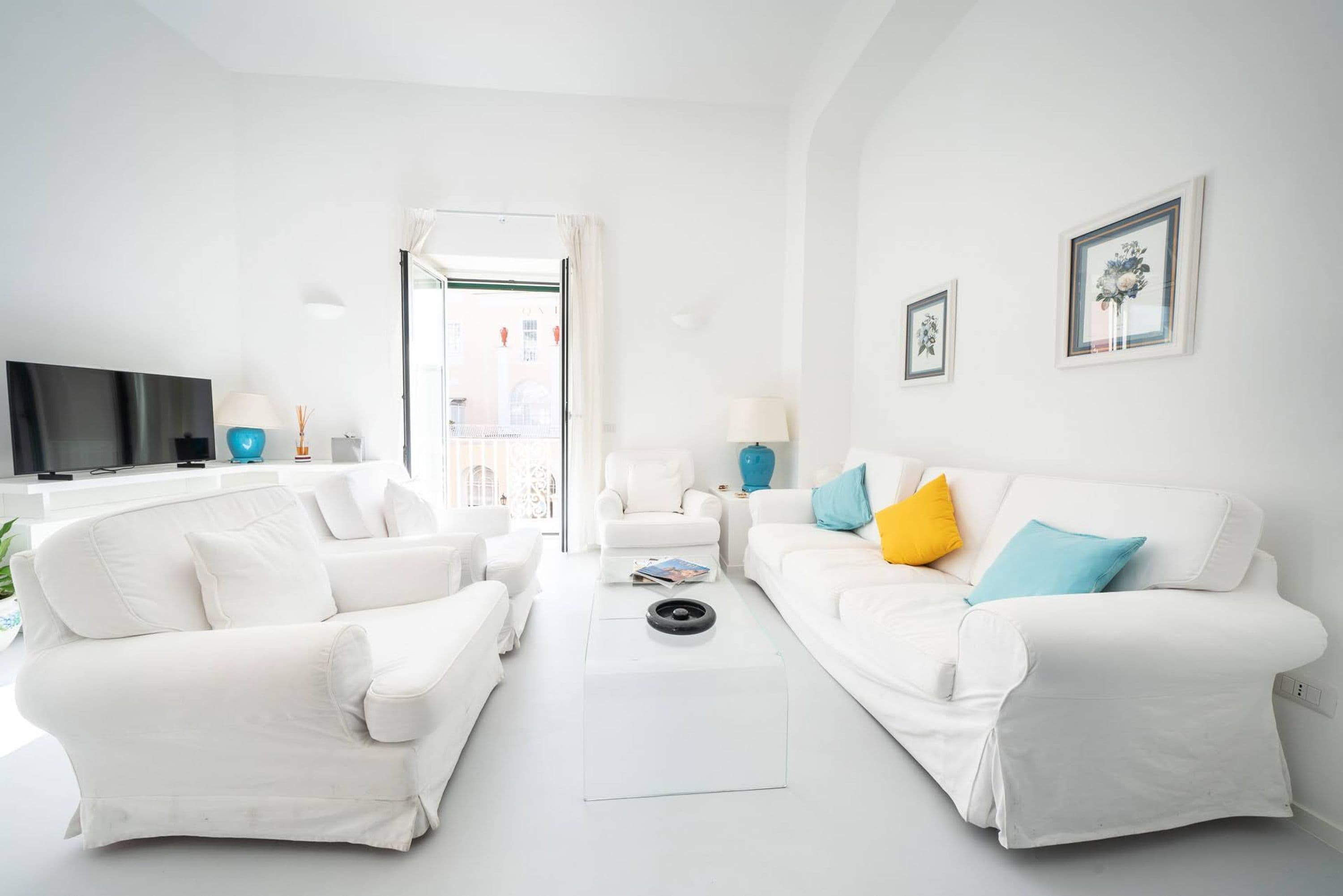 A modern living room with white furniture, a flat-screen TV, and a view through an open door.