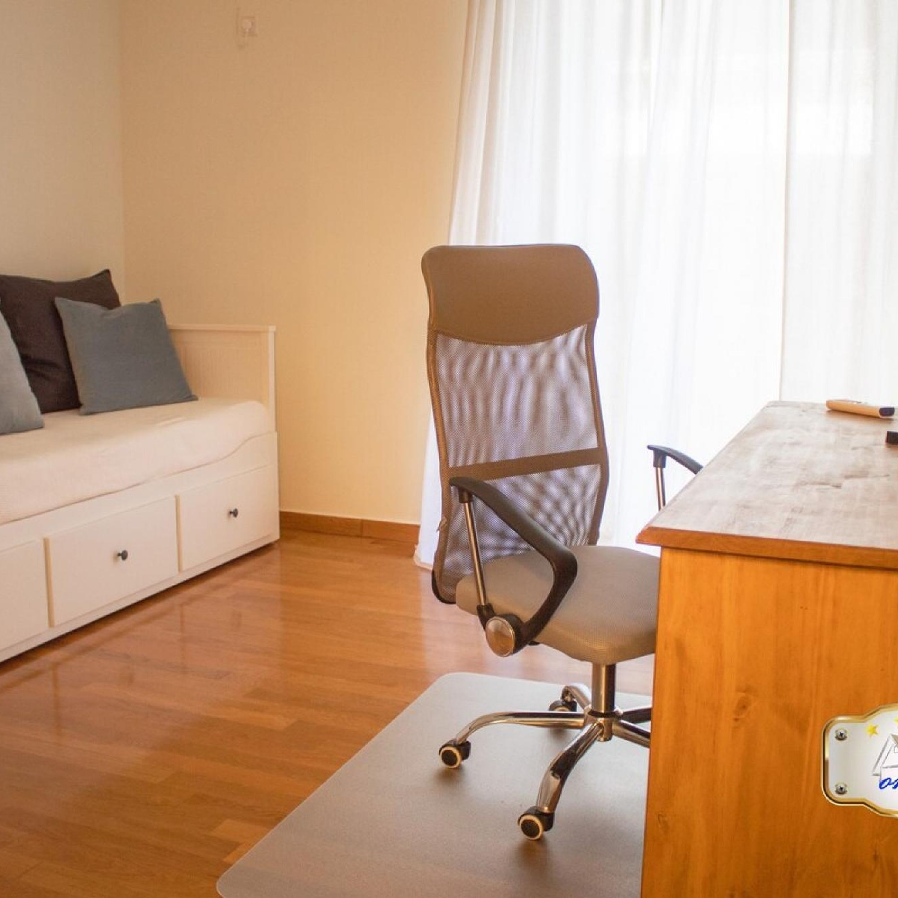 A room with a wooden desk, a cushioned chair, a white sofa with pillows, and a wooden floor.