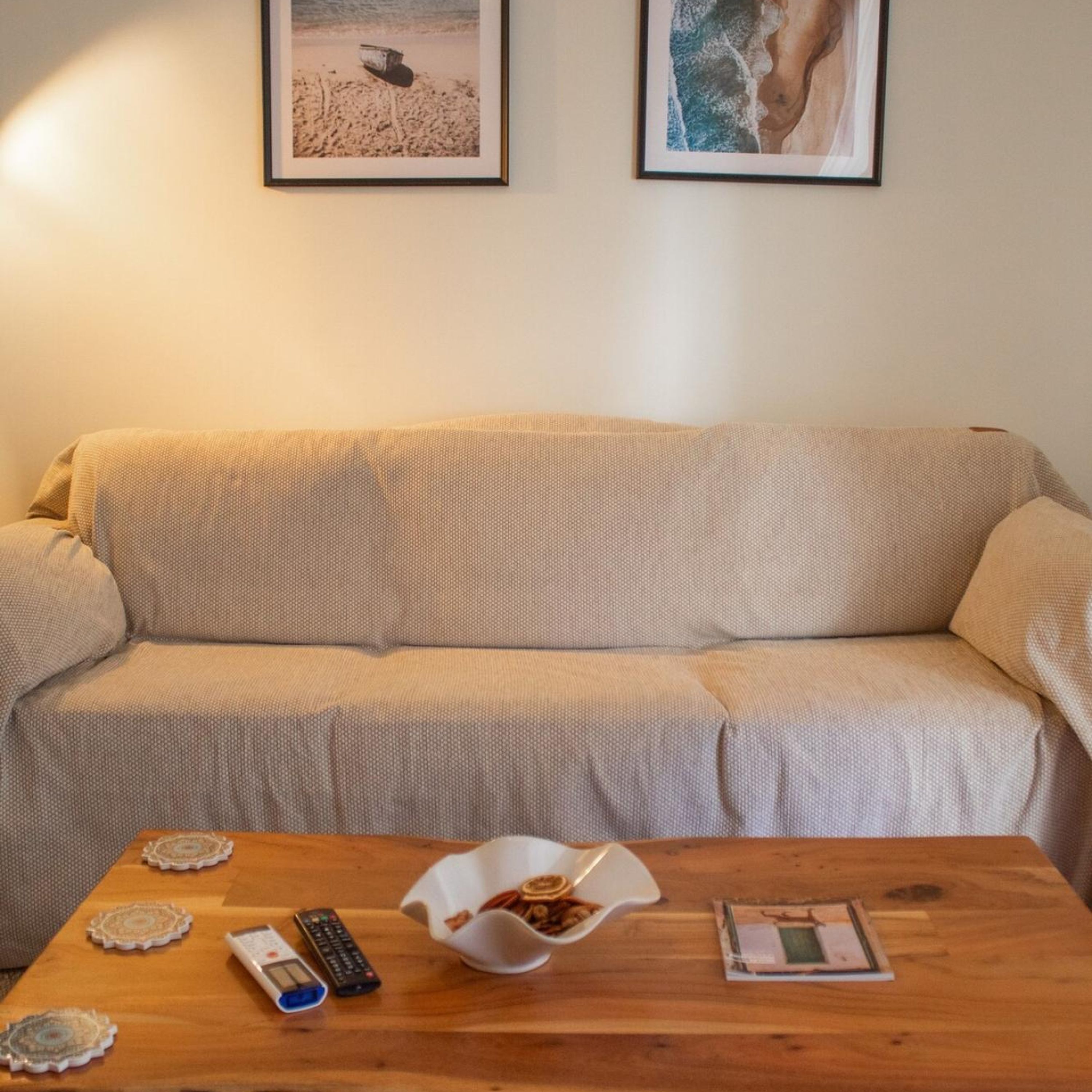 A living room with a beige sofa, a wooden coffee table with a bowl of snacks, a remote control, and decorative coasters.