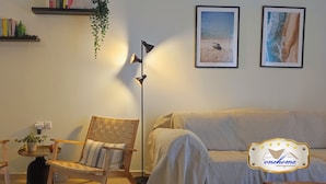 A cozy living room with a sofa, two wooden chairs, a floor lamp, and wall-mounted shelves with books and a hanging plant. - Filoxenia in the Centre of Kalamata (Kalamata)