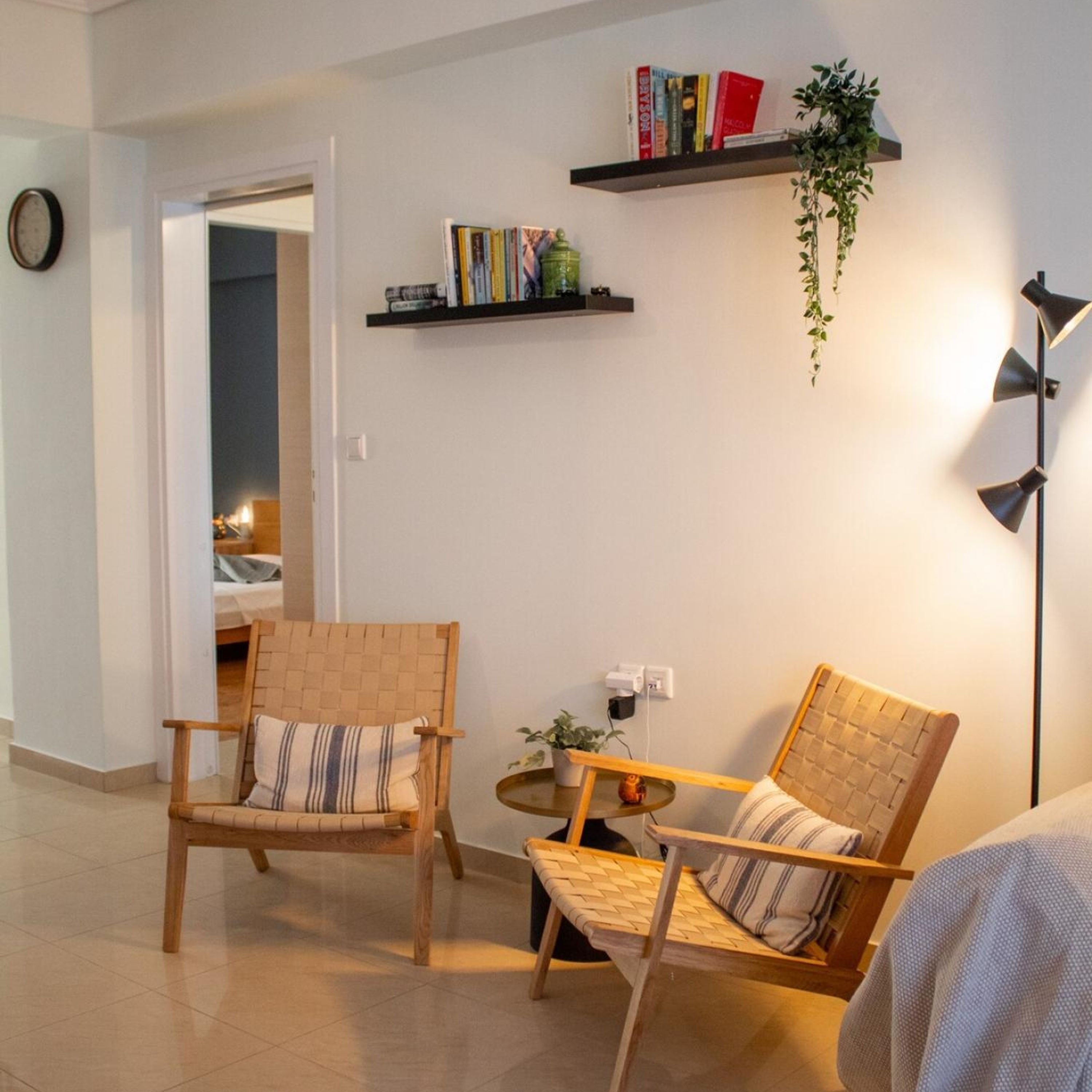 Un salon avec deux fauteuils en osier, une petite table d’appoint et un lampadaire. On y trouve deux étagères remplies de livres et une plante.