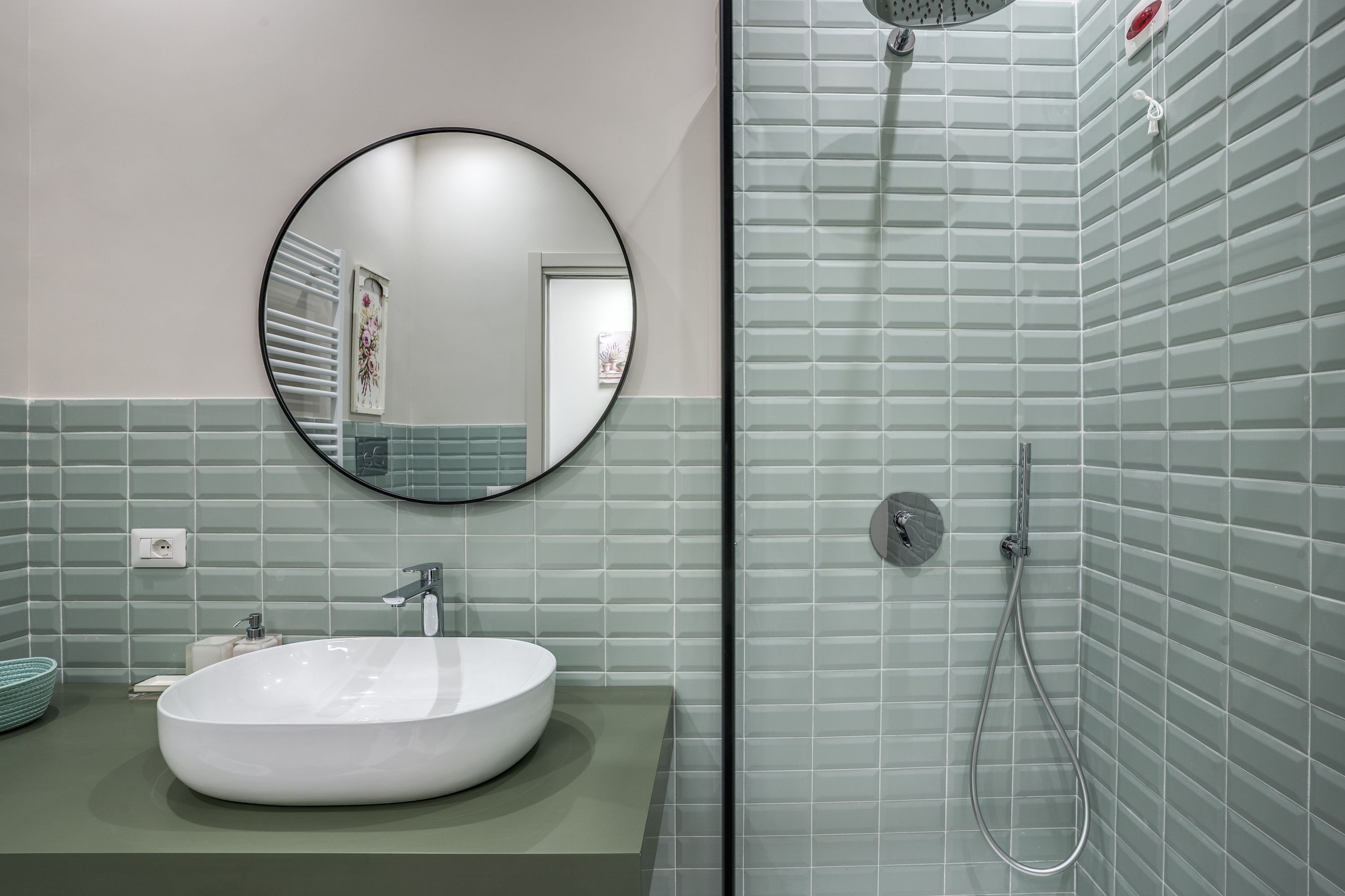 A bathroom with a round mirror, a white sink, and a shower area with tiled walls.