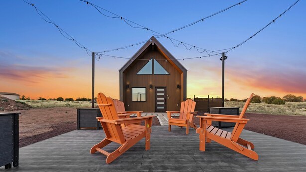Une cabane en bois avec un toit triangulaire, une véranda dotée de quatre fauteuils Adirondack et une guirlande lumineuse suspendue au-dessus.