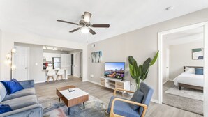 Una sala moderna con sofá, sillón, mesa de centro y televisor. Hay un comedor con sillas y mesa, y un dormitorio con cama y mesita de noche.
