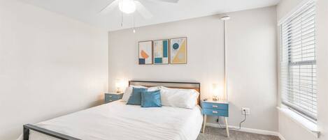 Un dormitorio con cama, ventilador de techo, lámparas de noche, una ventana con persianas y decoración abstracta en la pared.