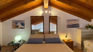 Un dormitorio con techo de madera inclinado, una cama con ropa de cama floral, dos espejos en la pared, una mesita de noche con lámpara, y una pequeña mesa con lámpara y caja de pañuelos.