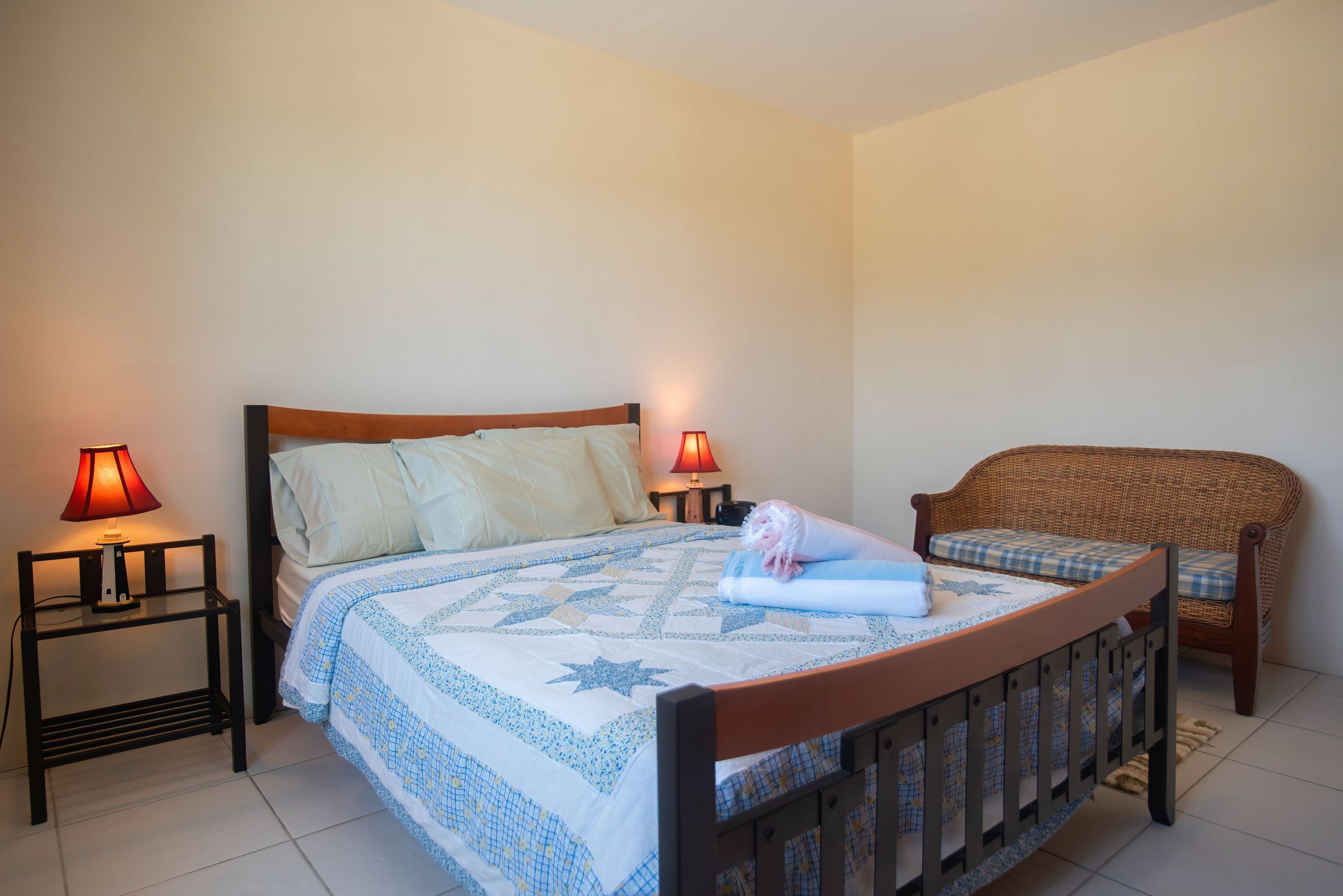 Quarto com cama, duas mesas de cabeceira com abajures, uma cadeira de vime e uma cama bem arrumada com um cobertor estampado com estrelas.