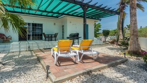 A covered patio with two yellow lounge chairs, a grill, and a pergola with a green roof. - Beautiful Luxury Home in Seroe Colorado (San Nicolas Zuid)