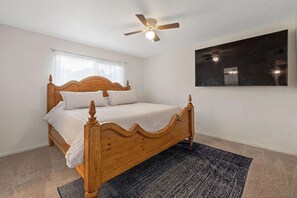 Un dormitorio con cama de madera, ventilador de techo, espejo grande y ventana con persianas.