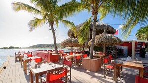 An outdoor dining area with wooden tables and red chairs, surrounded by palm trees and thatched umbrellas. - Artiko Luxury Suites - Jan Thiel (Jan Thiel)