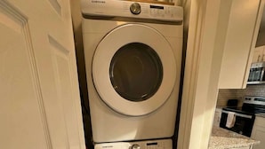 A compact laundry room with stacked washers and dryers, a small shelf with toiletries, and a kitchen area visible through an open door. - Modern Cozy Walk up With King Bed (Wilmington)