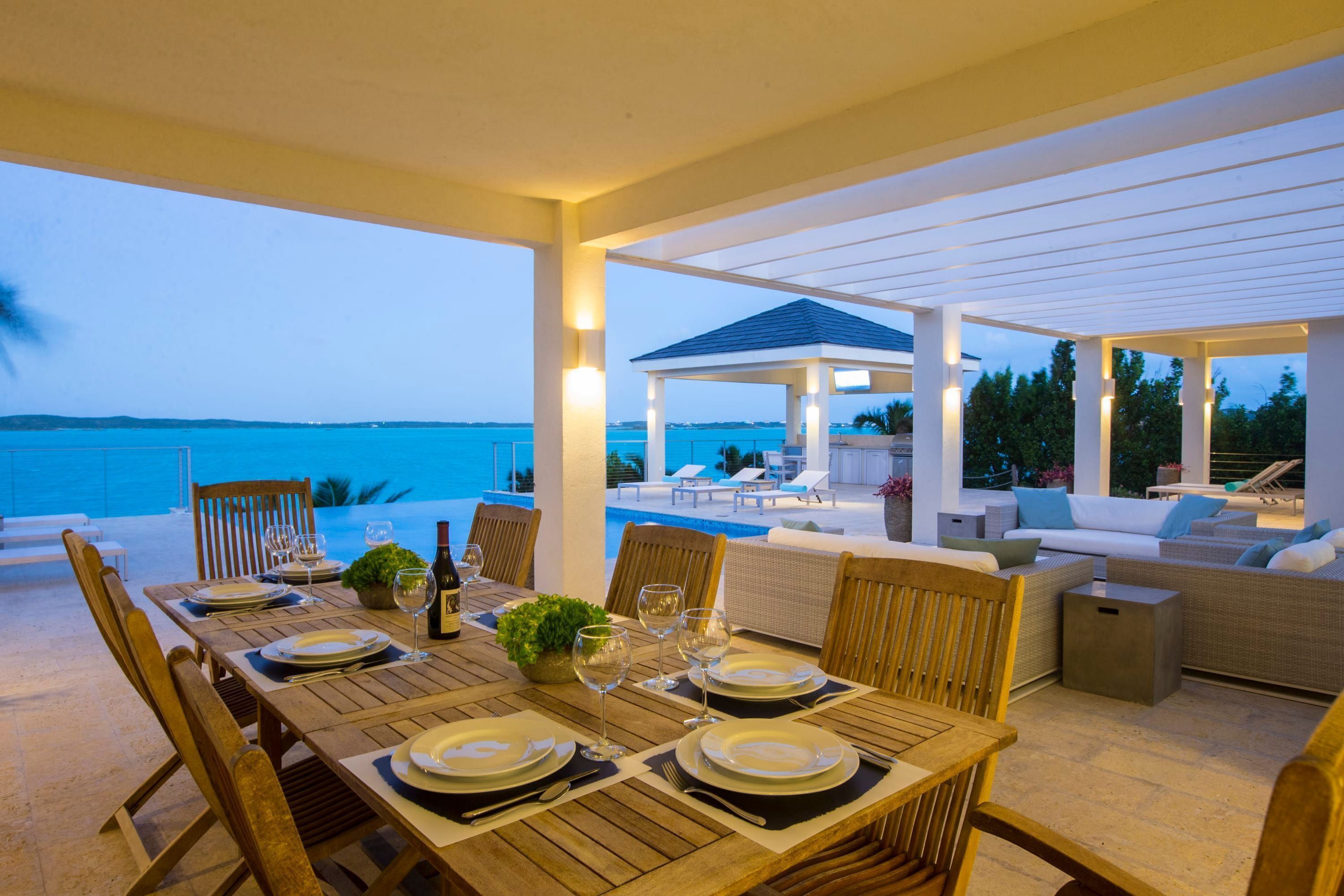 An outdoor dining area with a view of the sea, wooden tables and chairs, and a covered seating area with cushions.