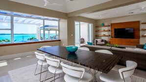 A modern living room with a large dining table, white chairs, a TV mounted on a wooden wall, and a view of the ocean through large windows. - 5 BR Waterfront Villa On Chalk Sound Blue Water (Providenciales)