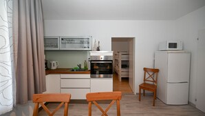 Uma cozinha compacta com armários brancos, micro-ondas e geladeira. Há uma mesa de jantar de madeira com duas cadeiras.