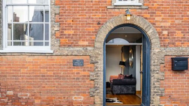A brick building with a large arched doorway leading to a room with a bed, a lamp, and a small table. - Host Stay The Chapel in Thornham (Hunstanton)