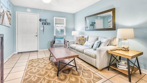 A living room with a light blue wall, a white door, a ceiling fan, a wooden coffee table, a striped sofa, a patterned rug, and a side table with a lamp. - Sunrise Village 215- 2BR 1BA Gulf Front Sleep 6 (Gulf Shores)