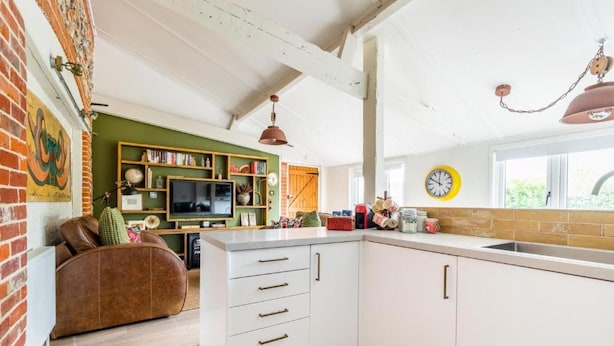 A kitchen with white cabinets, a brown leather sofa, and a green wall with a clock. - Host Stay Nicholson Cottage (North Walsham)
