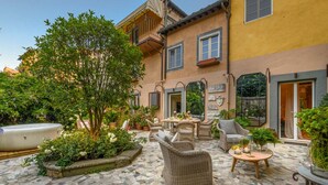 A courtyard with a stone floor, wicker furniture, and a large tree. - The Garden Lodge in Lucca (Lucca)