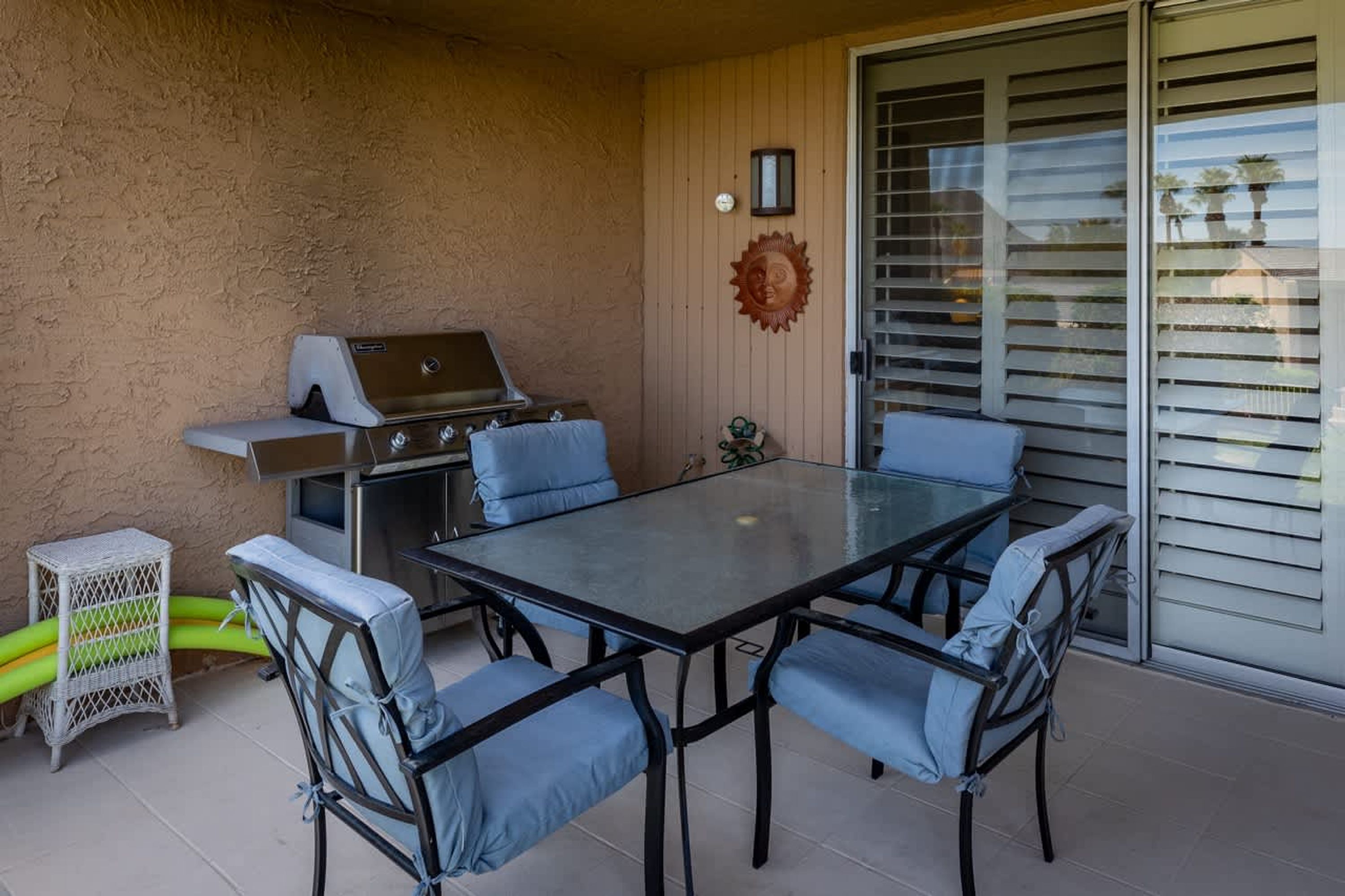 An outdoor patio with a glass-top table, six blue cushioned chairs, a barbecue grill, and a wicker stool.