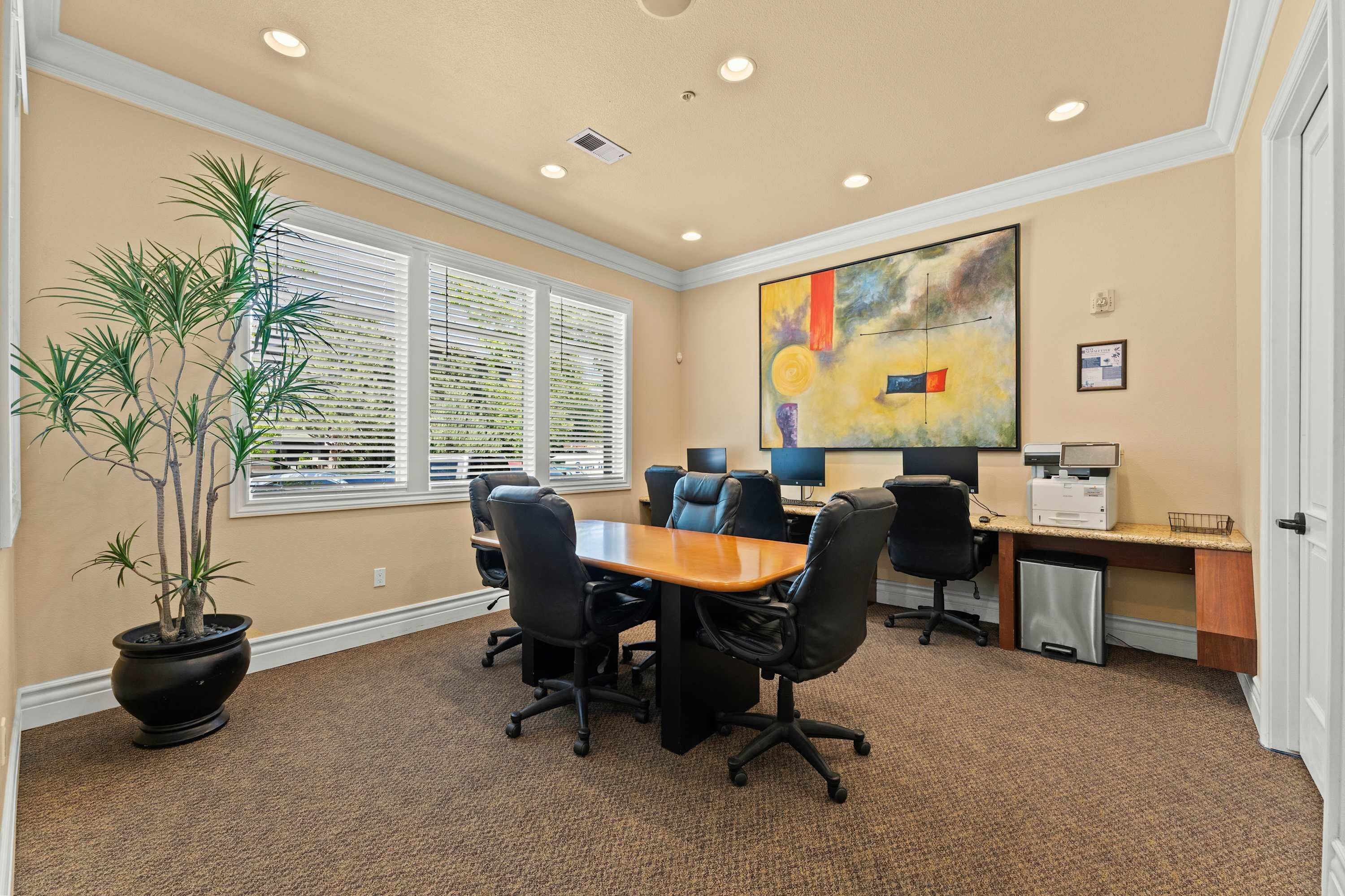 A conference room with a wooden table, black office chairs, a potted plant, and a large abstract painting on the wall.