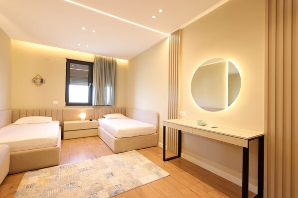A modern hotel room with two single beds, a bedside table with a lamp, a round mirror, and a window with curtains. - Grandstay Apartments 006 (Durrës)