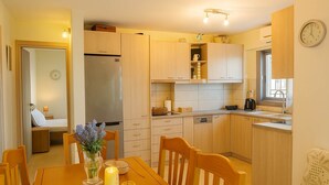 Cocina compacta con armarios de madera, un refrigerador, una mesa de comedor con sillas y un reloj en la pared.