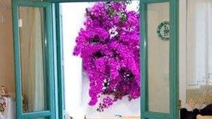 A dining area with a table set for two, a chair, and a view of a balcony with purple flowers. - Beachside Bliss at Skyros Luxury Retreat (Skiros)