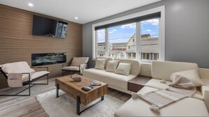 Ein modernes Wohnzimmer mit einer Couch, einem Sessel und einem Couchtisch. Ein Flachbildfernseher ist an der Wand befestigt, und ein Fenster bietet Blick auf Gebäude im Freien.