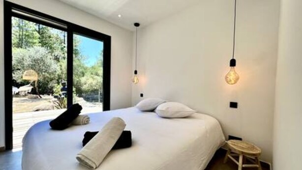 A modern bedroom with a large bed, a small stool, and a view of the outdoors through a large window. - Casa di Macchjerolu - 8 Pers (Sotta)