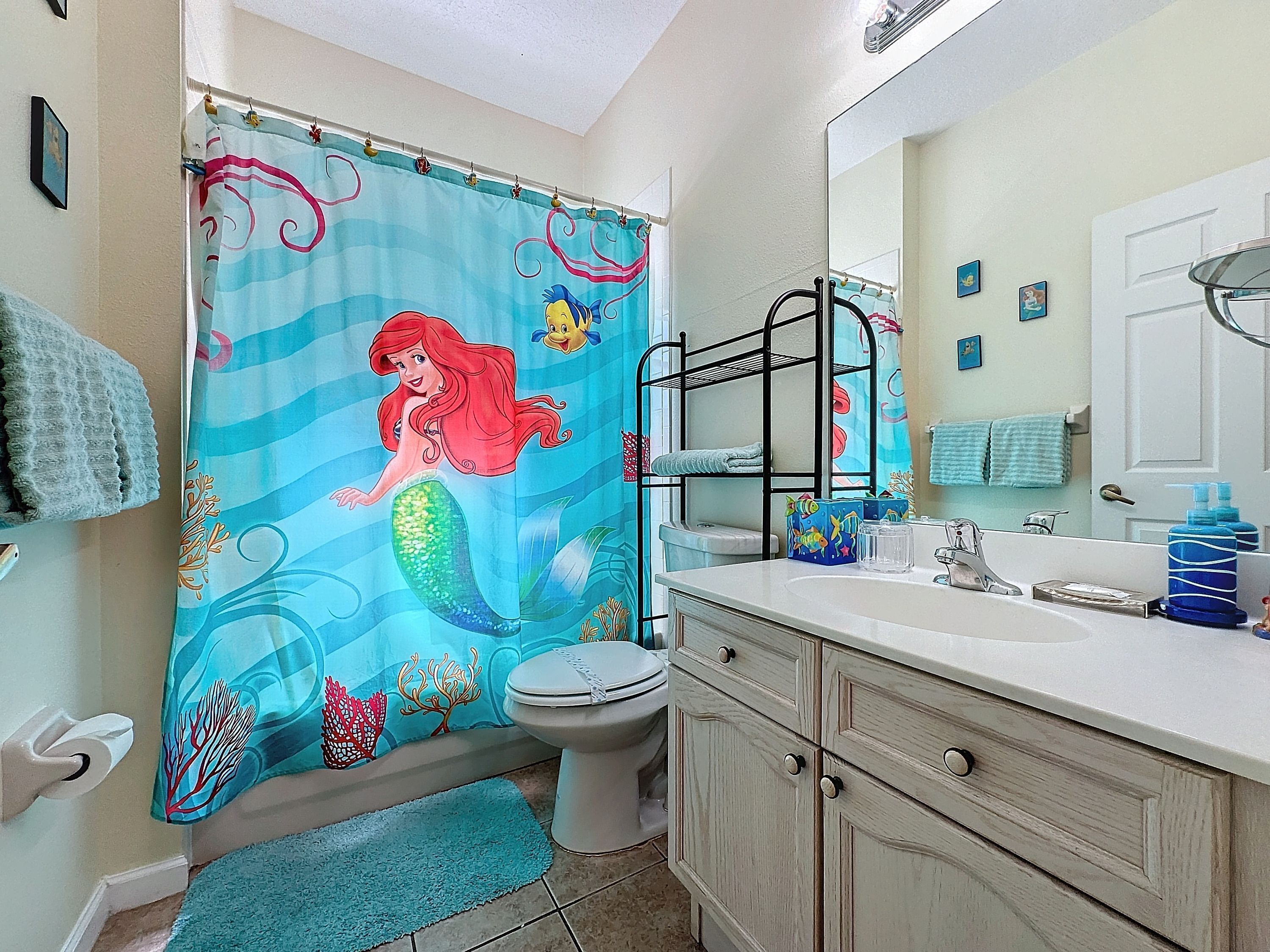 A bathroom with a mermaid shower curtain, a toilet, a sink with a mirror, and a vanity with drawers.