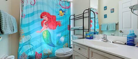 A bathroom with a mermaid shower curtain, a toilet, a sink with a mirror, and a vanity with drawers.