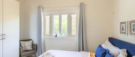 A bedroom with a bed, a chair, a window with curtains, and a wall with framed pictures.