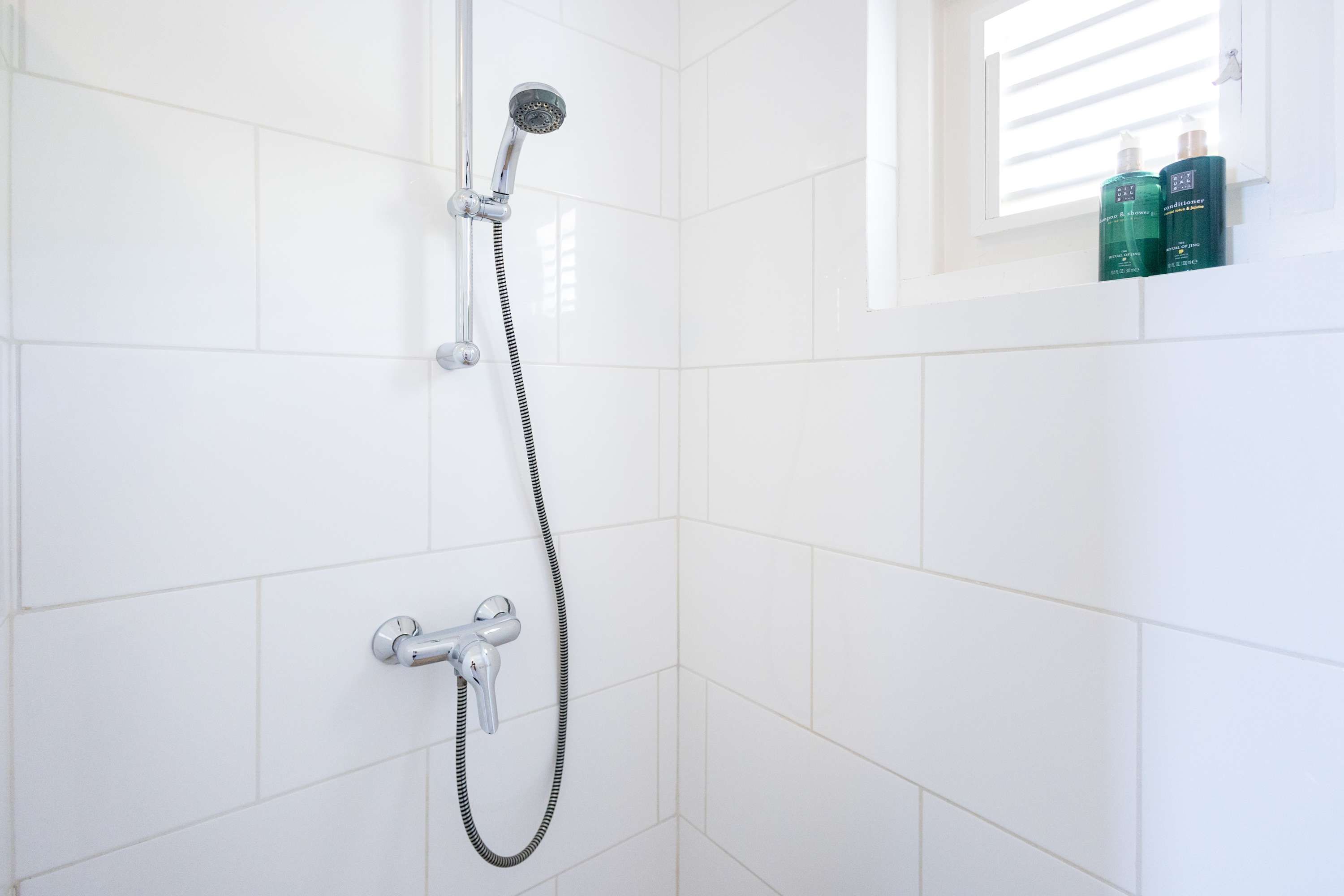 A shower with a handheld showerhead and a wall-mounted faucet. There are two bottles on a shelf above the shower.