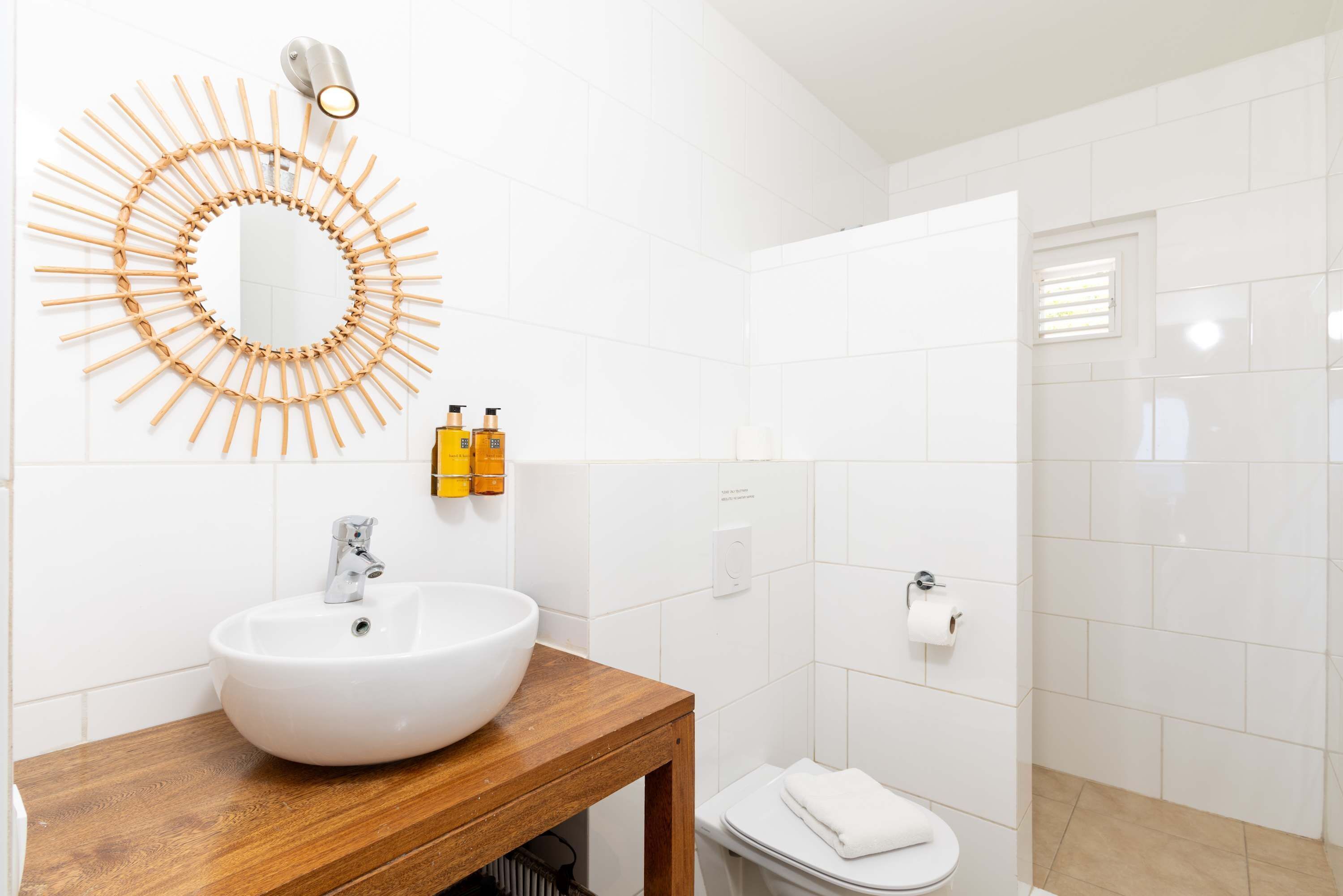 Et badeværelse med en trækommode, en hvid vask, et spejl med solstråle-design og et toilet.