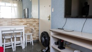 Una habitación con un escritorio blanco, dos taburetes blancos, un ventilador negro, un televisor montado y un teléfono en una estantería.