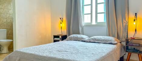 A bedroom with a bed, a nightstand, a lamp, a window with curtains, and a toilet.
