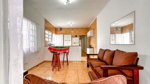 A living room with a brown sofa, a coffee table with a red and green tablecloth, and a kitchen in the background. - Tima Casa com Wifi pr Ximo a Praia em Itanha m SP (Itanhaém)