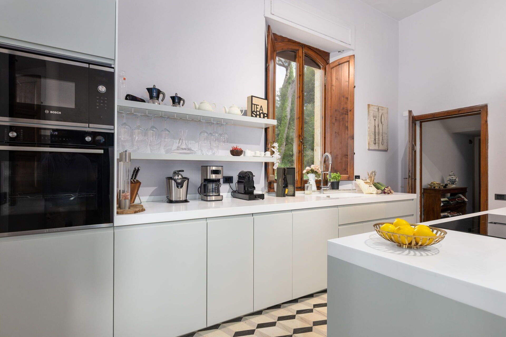 A modern kitchen with white cabinetry, a Bosch oven, a coffee machine, and a window with a view of trees.