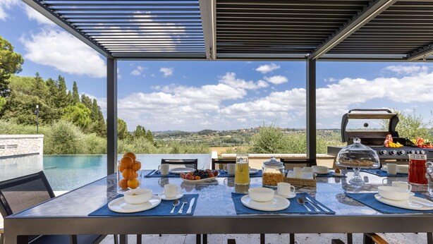 An outdoor dining area with a view of the countryside, equipped with a grill and a table set for breakfast.