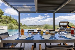 An outdoor dining area with a view of the countryside, equipped with a grill and a table set for breakfast.