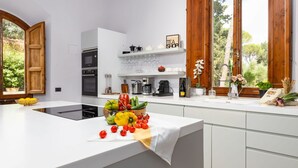 A modern kitchen with white cabinetry, a central island with fresh produce, and a view of greenery through large windows.